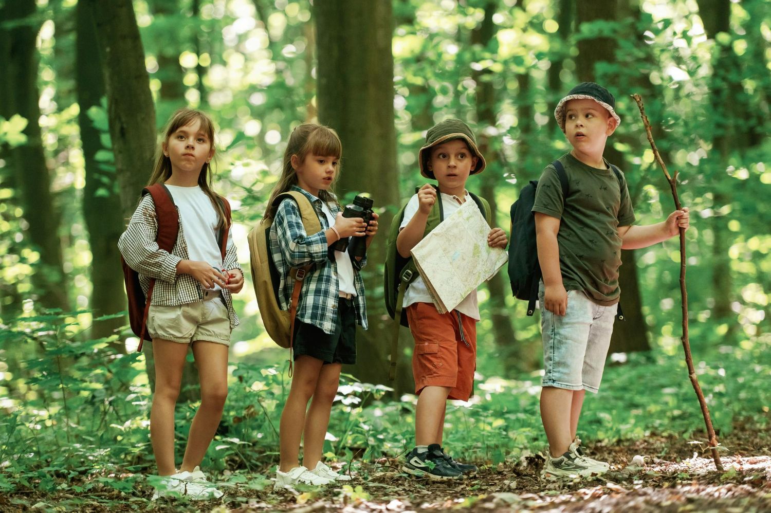 4 children on a hike in the woods at summer camp with hats, backpacks, map, and a walking stick
