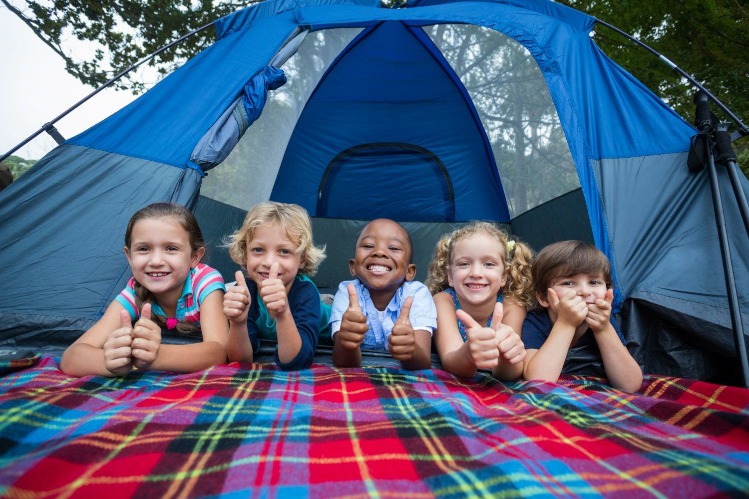 5 school age kids lying on their tummies on a plaid blanket in front of their tent at summer camp, all smilies and giving two thumps up