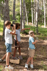 three elementary age children exploring the woods at summer camp with binoculars