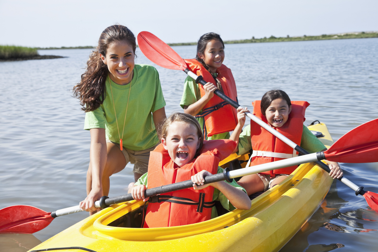 Summer camp counselor with kids at waters edge and kayak, everyone is laughing