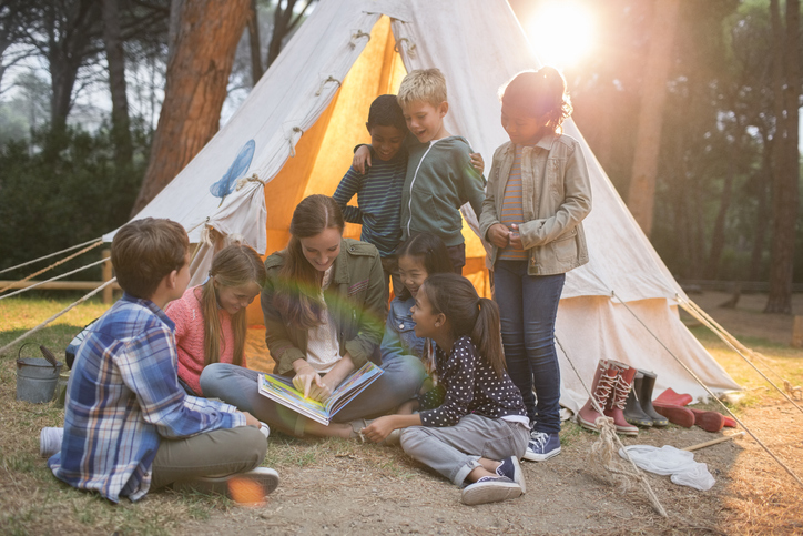 camp counselor and kids reading a book in front of pitched tent at summer camp