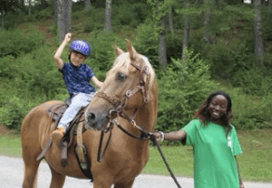 Young girl riding horse with arm in the air being lead by camp consoler