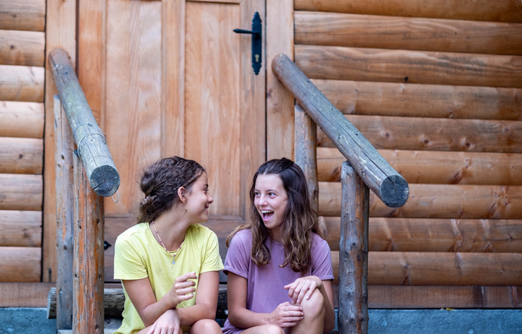 two teen girls are sitting in front of a camp cabin talking, laughing and joking