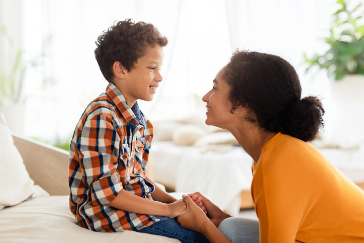Joyful mother and son engaging in chat about summer plans, talking at home, sitting holding hands and smiling to each other. Trust and deep family bond, mother and kid relationship, communication. Side view shot