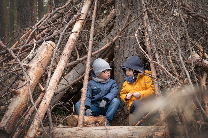 Two warmly dressed little boys in a winter pine forest play in a makeshift hut (self made branches shelter). Lifestyle. Outdoors child's games, recreation at forest.