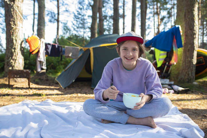 An 11 year old boy or teenager has breakfast with cereal and milk, sitting on a blanket in front of a tent in the forest, spending summer holidays in a tent camp.