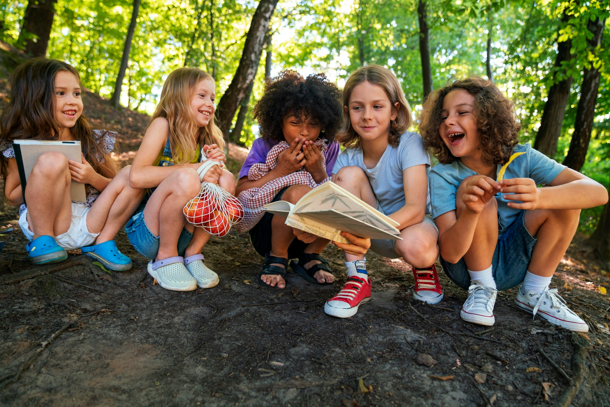 5 elementary age camps perched on a wooded hilltop studying a nature field guide and laughing