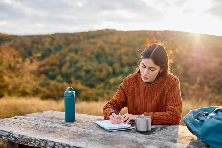 Woman writing notes in her journal sitting at a wooden table outdoors, enjoying a tranquil moment in autumn nature
